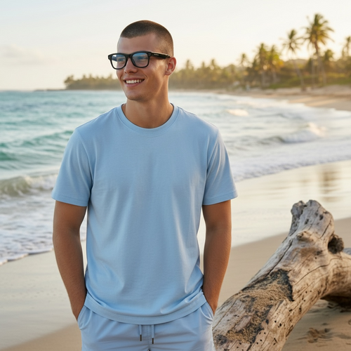 Smiling man wearing VIZER VISION APEX glasses and a light blue t-shirt standing on a beach with waves and palm trees in the background.
